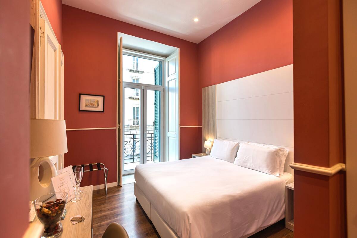 Una camera d'albergo con un letto, una scrivania con una lampada, una sedia e vista all'esterno.