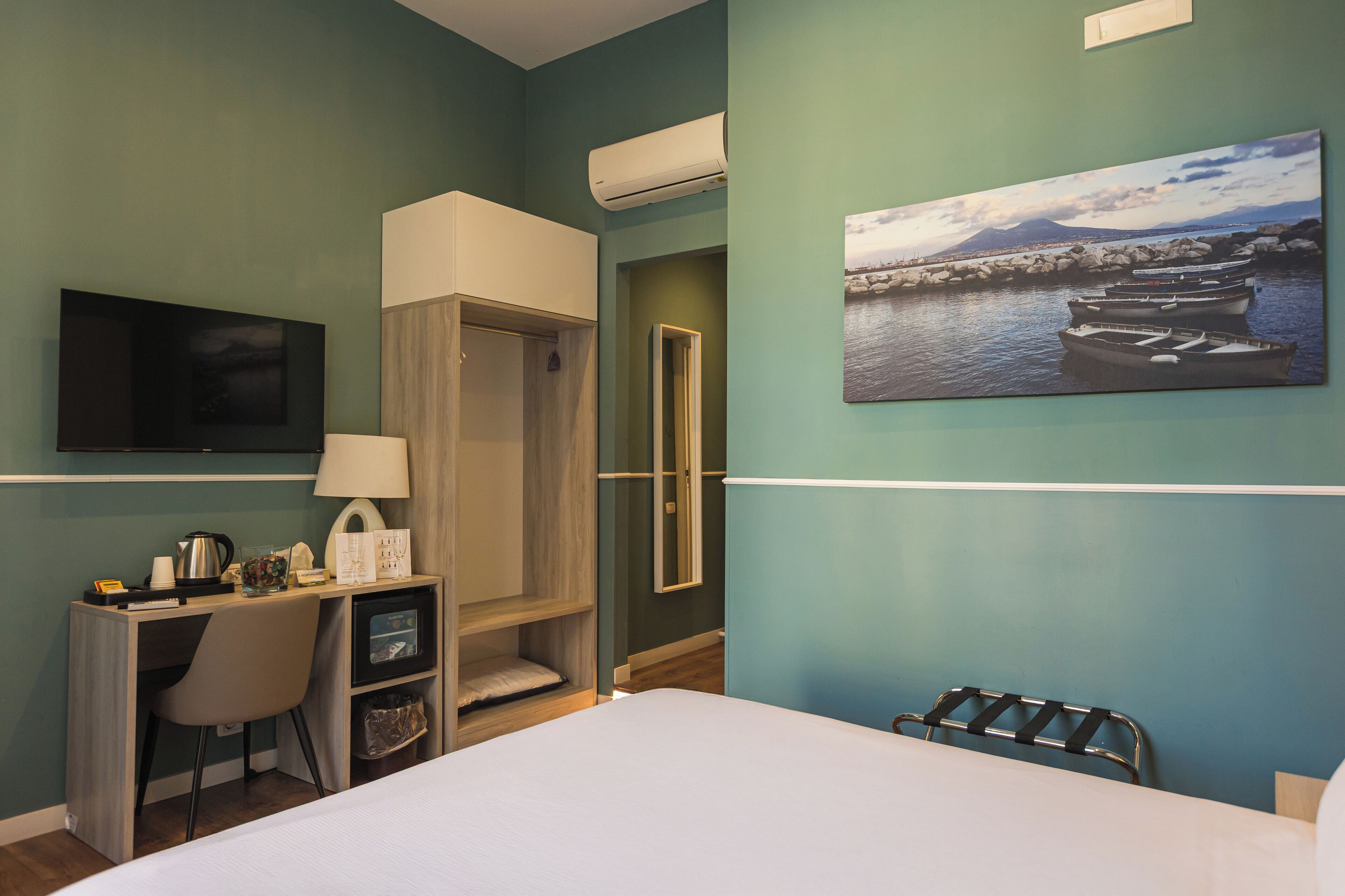 A hotel room with a bed, a desk with a chair, a TV, an air conditioner, a painting, and a wardrobe.