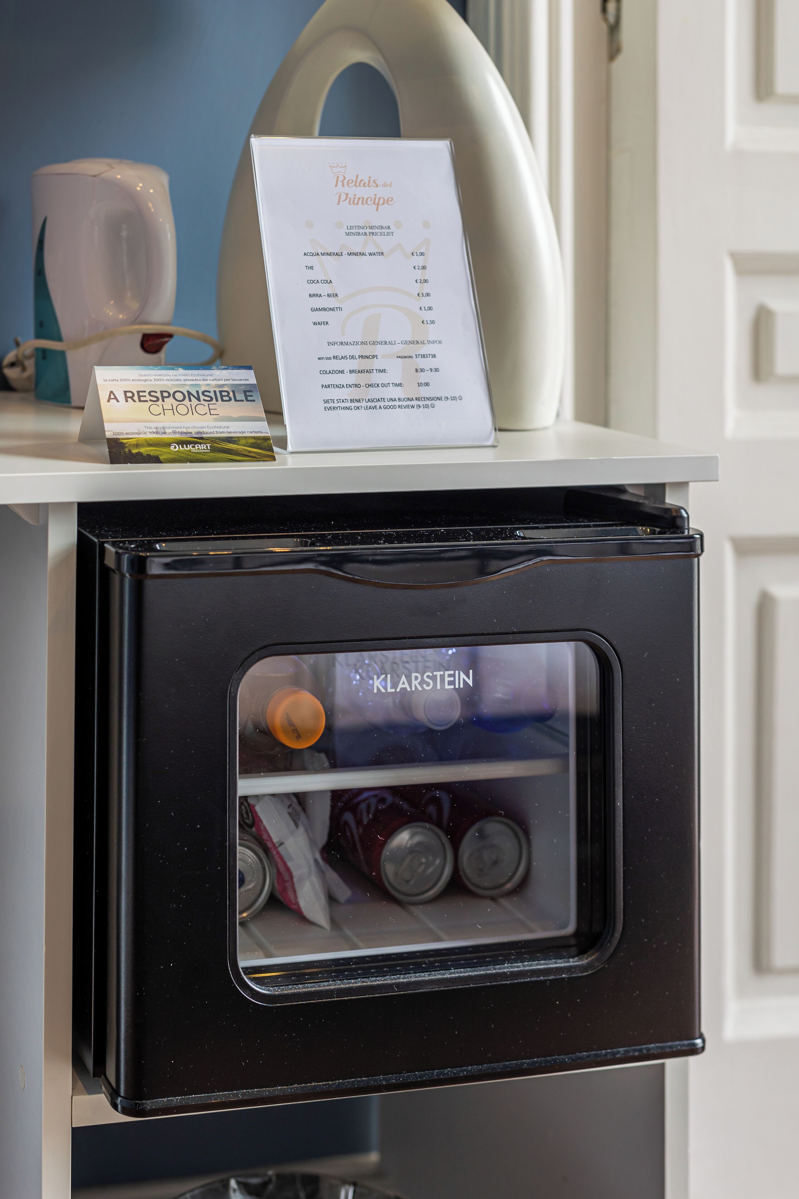 A black minibar with a glass door, containing cans and a bottle. A menu titled "Relais del Pincio" is visible on the counter above.
