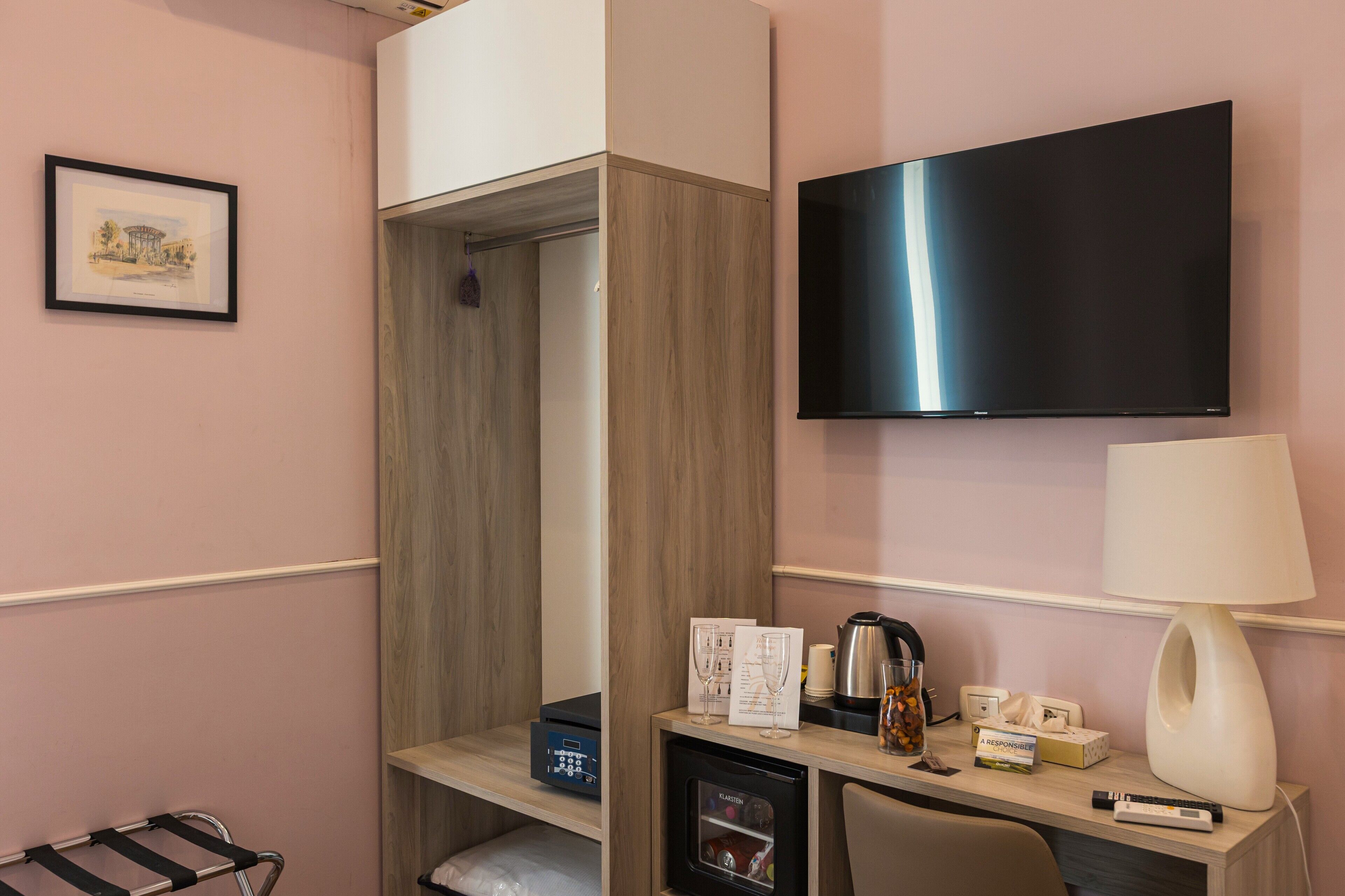 A hotel room with a wooden wardrobe, a flat-screen TV, a desk with a lamp, a coffee maker, and a minibar.