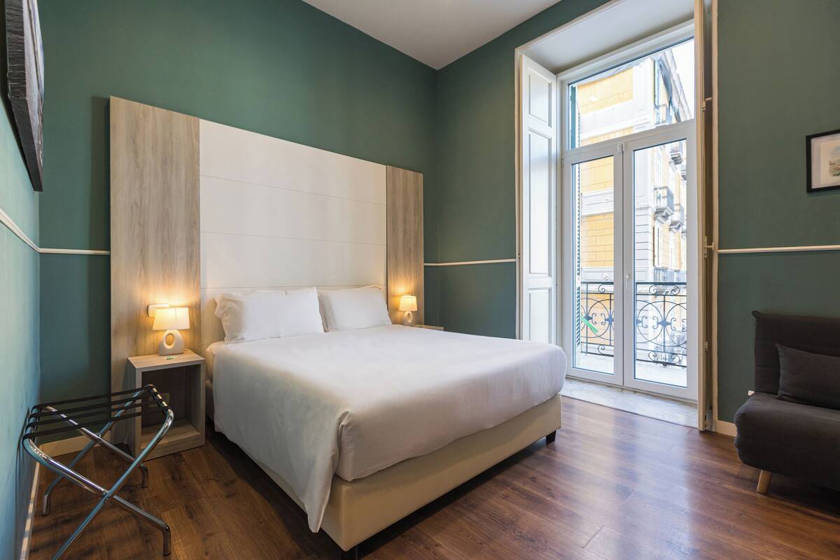 Un letto rifatto con cura in una stanza con un'ampia finestra, un comodino con una lampada e una sedia.
