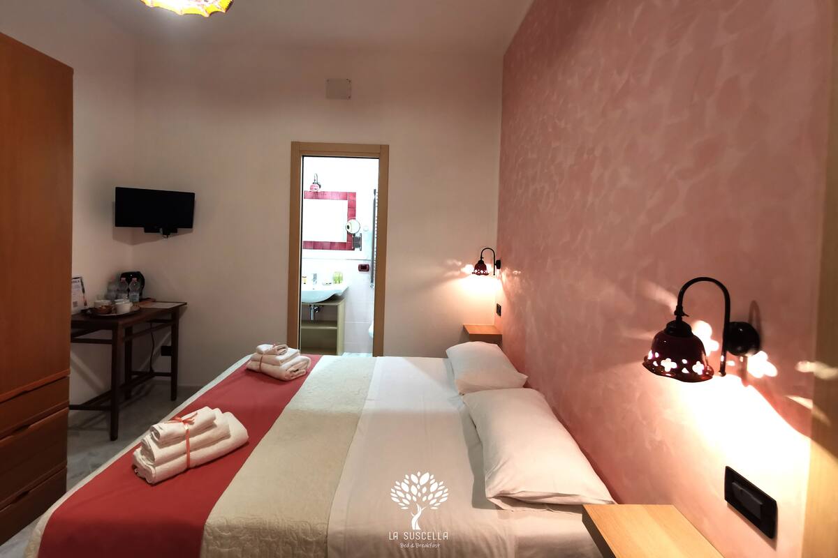 A hotel room with a bed, bedside table, wall-mounted TV, and a bathroom visible through an open door.