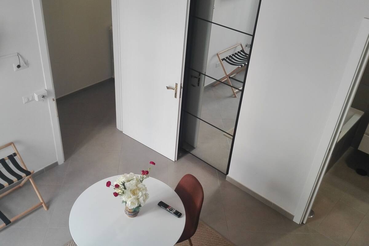 A modern room with a white door, a small round table with a vase of flowers, a remote control, and a striped bench.