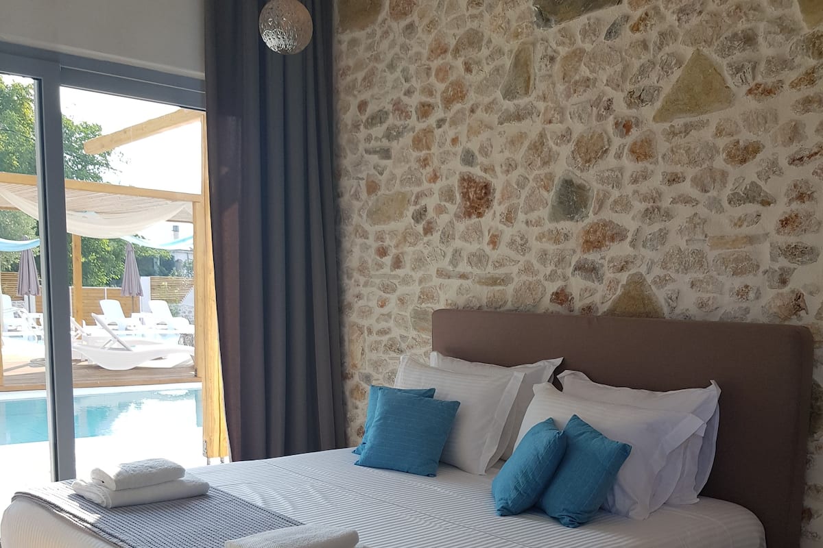 Ein Schlafzimmer mit Steinwand, einem Bett mit weißer und blauer Bettwäsche sowie Blick auf einen Pool.