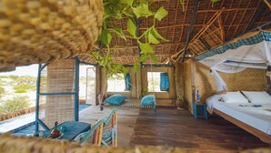 A room with a bed, a hanging chair, a small table, and a window with curtains. - Kizingo Beach Eco Lodge (Lamu)