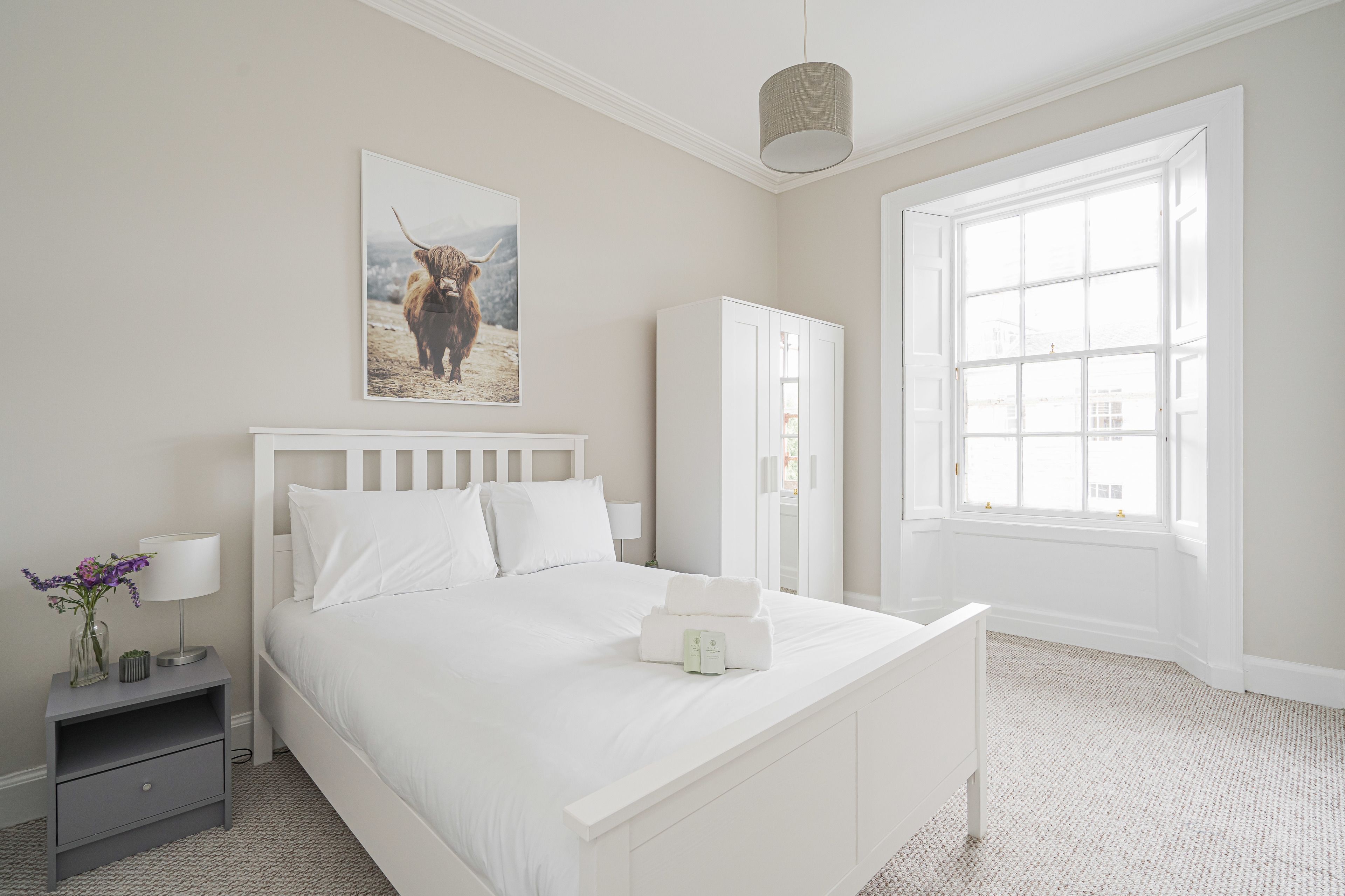 A bedroom with a bed, a nightstand, a wardrobe, a window, and a wall-mounted lamp.
