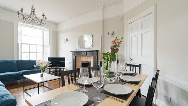 A dining area with a wooden table set for four, surrounded by black chairs. A blue sofa and a small coffee table with a vase of flowers are visible in the foreground. - Warriston Crescent Apartment (Edinburgh)