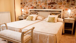Una cama doble con cabecera de madera, dos mesitas de noche con lámparas, un banco y una pared de piedra.