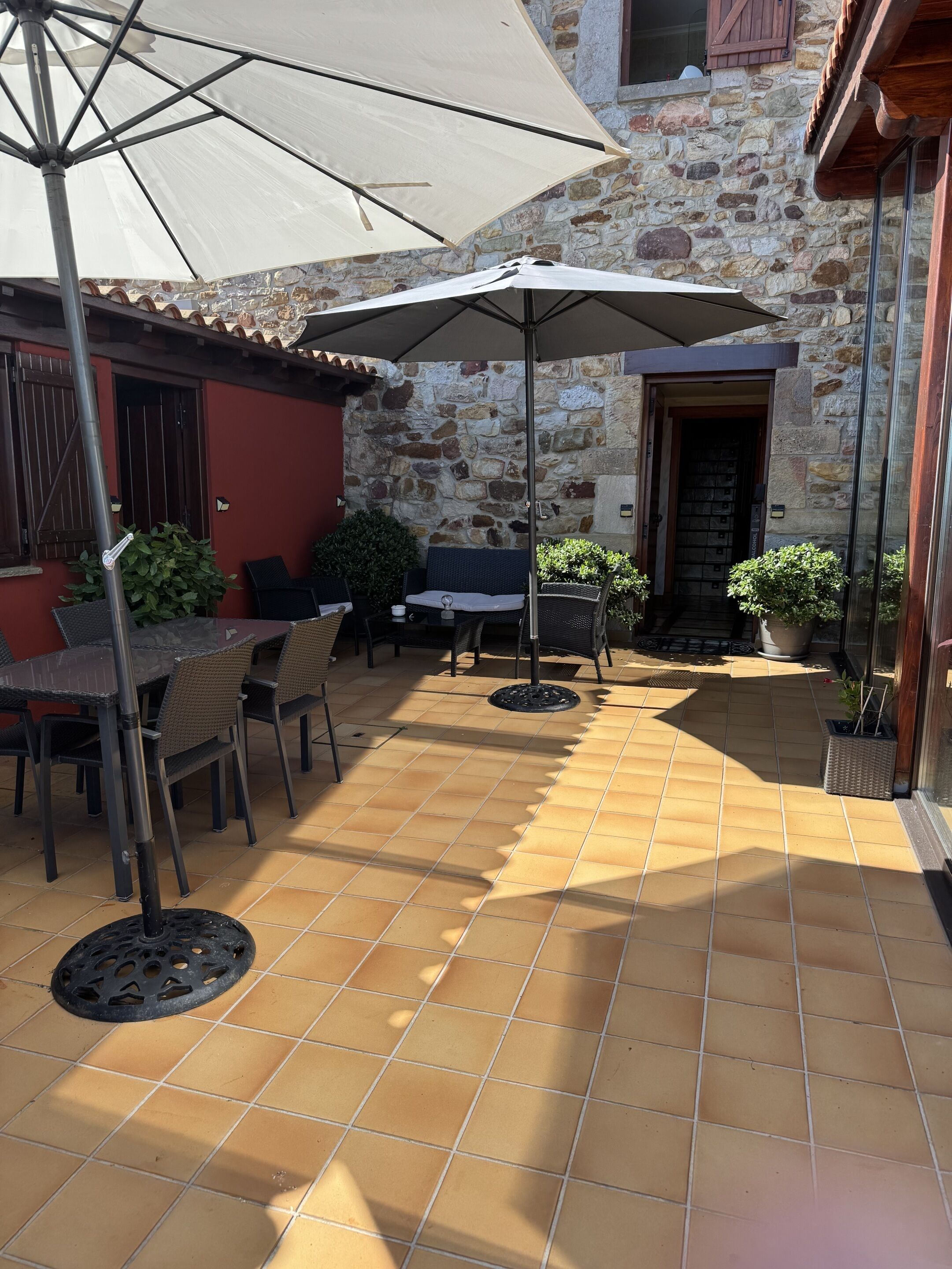 An outdoor patio with tiled flooring, a table with chairs, a large umbrella, and potted plants.