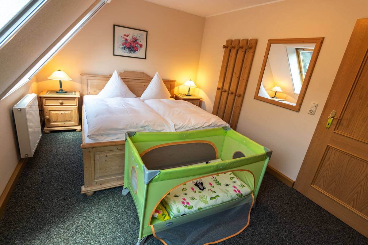 A bedroom with a bed, two bedside tables with lamps, a green baby crib, a wooden door, and a window with a view.
