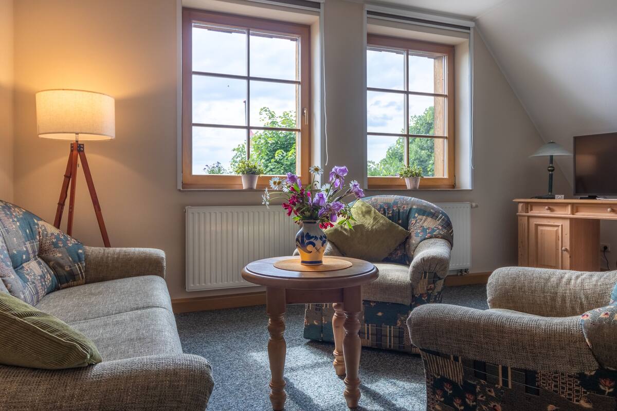 A cozy living room with a sofa, two armchairs, a small wooden table with a vase of flowers, a floor lamp, and a television on a wooden cabinet.