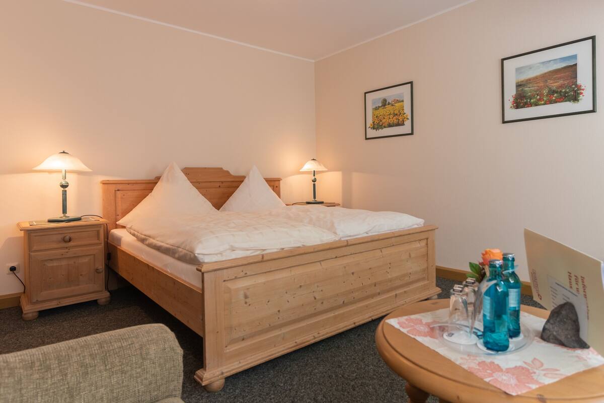 A wooden bed with white bedding, two bedside lamps, a small table with bottles, and framed pictures on the wall.
