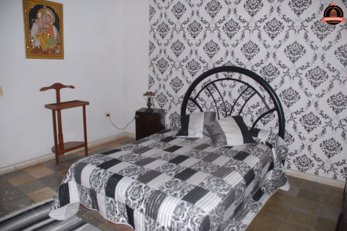 Une chambre à coucher comprenant un lit, une table de chevet, une chaise et un mur recouvert d’un papier peint à motifs.