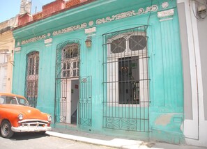 Un bâtiment de couleur turquoise, agrémenté de grilles en métal ouvragées, devant lequel est garée une voiture ancienne.
