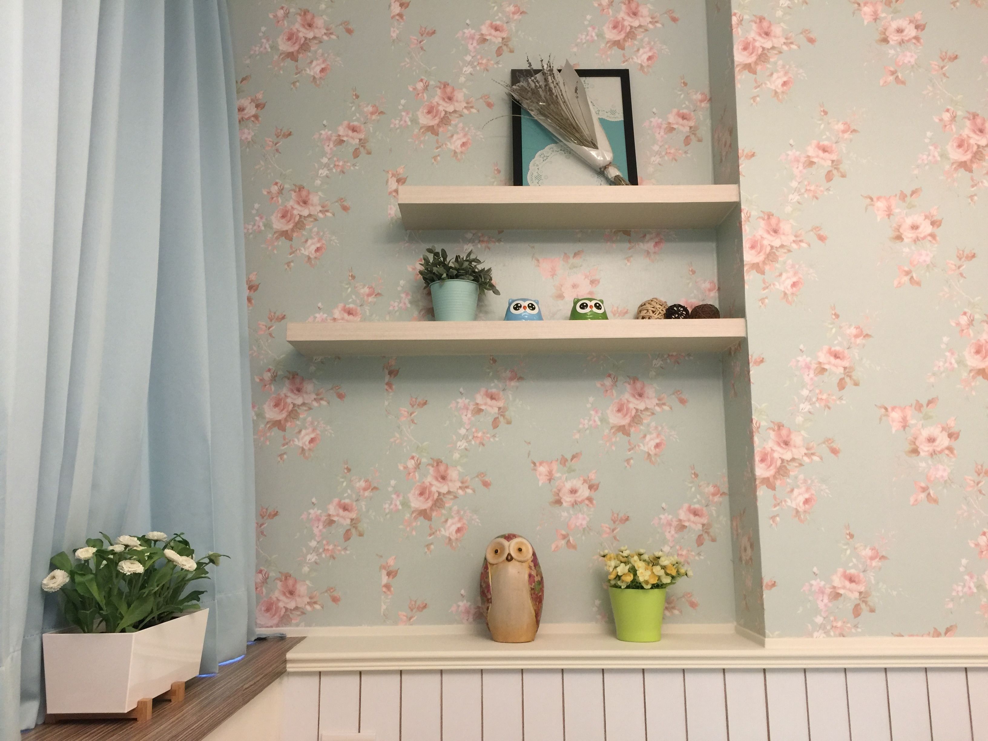 A room with floral wallpaper, a wooden shelf with decorative items, and a potted plant.