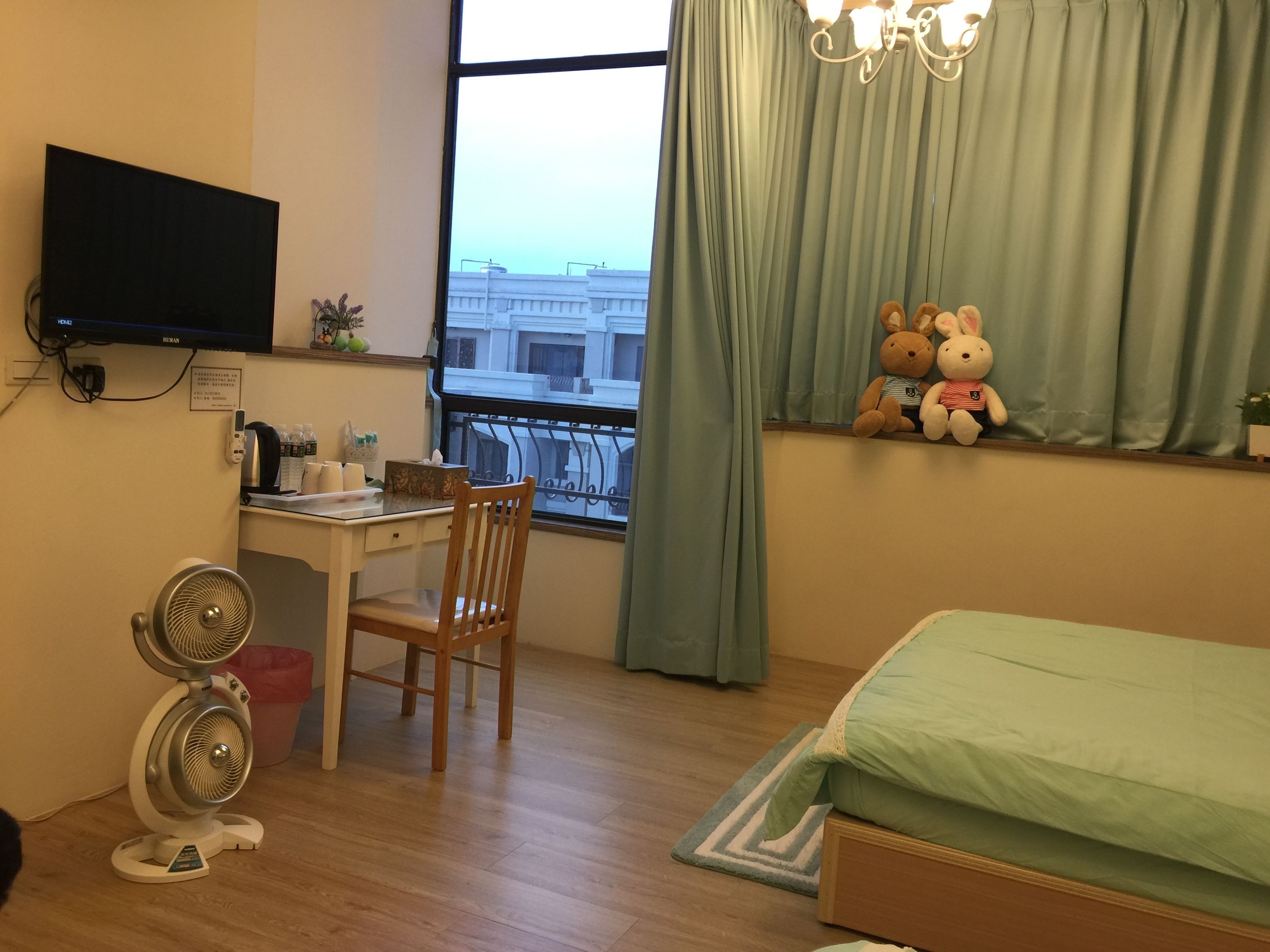 A room with a desk, chair, two fans, a bed, and a window with curtains.