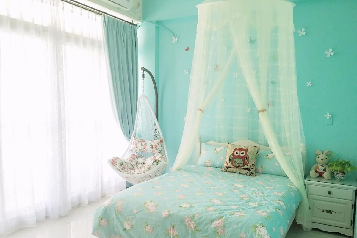 Een slaapkamer met een hemelbed, bloemenbeddengoed, een hangstoel en een nachtkastje met een teddybeer.