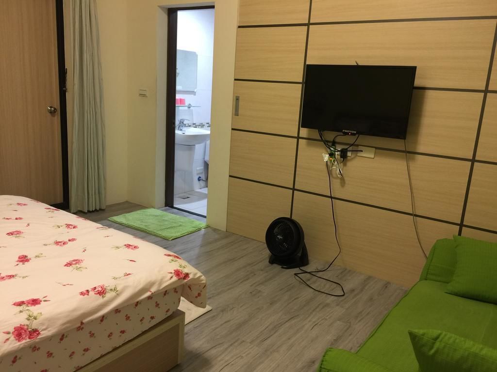 A hotel room with a bed, a green sofa, a flat-screen TV mounted on the wall, a fan on the floor, and a view of the bathroom through an open door.