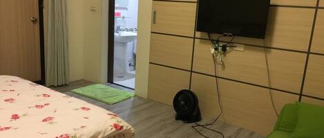 A hotel room with a bed, a green sofa, a flat-screen TV mounted on the wall, a fan on the floor, and a view of the bathroom through an open door.