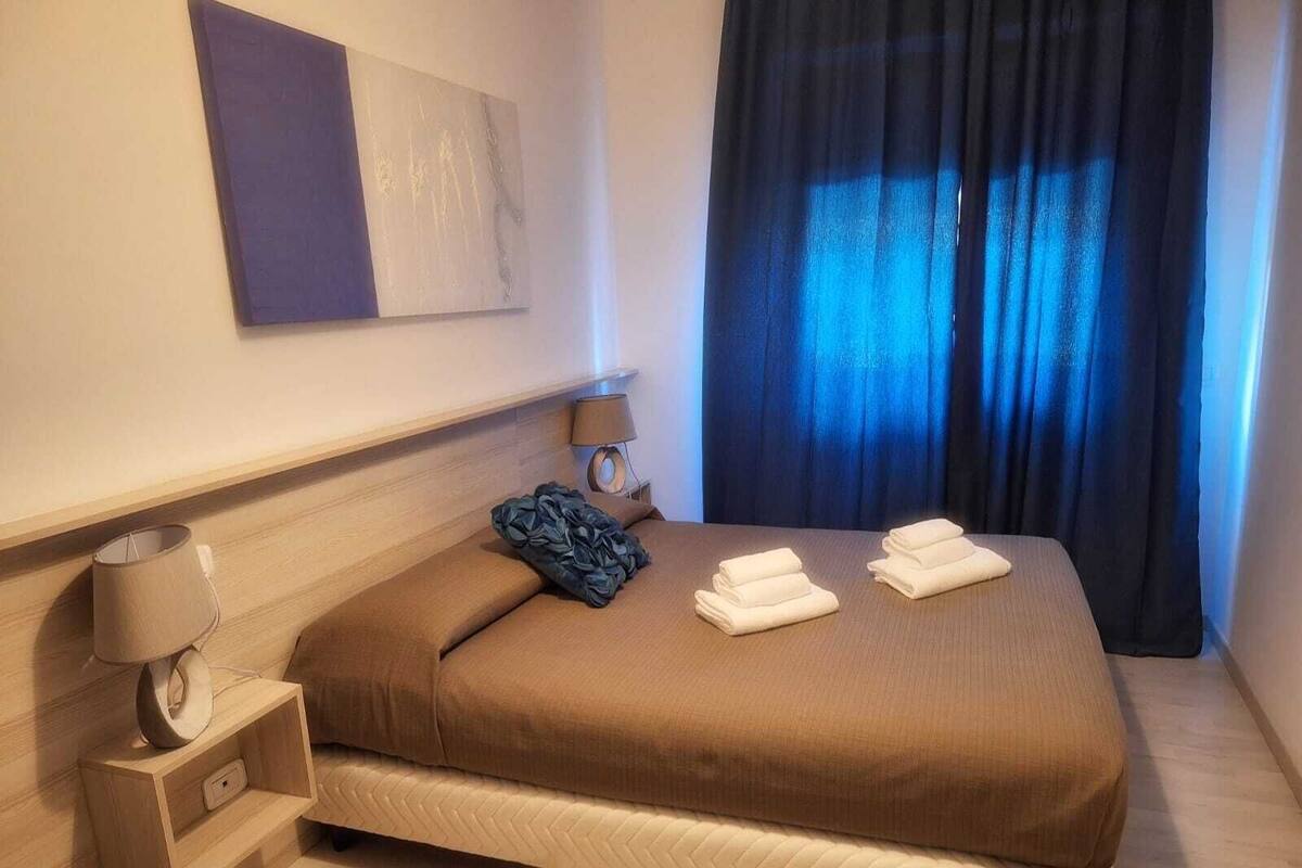 Ein Hotelzimmer mit einem Bett, einem Nachttisch mit Lampe, einem Wandbild und einem Fenster mit Verdunkelungsvorhängen.