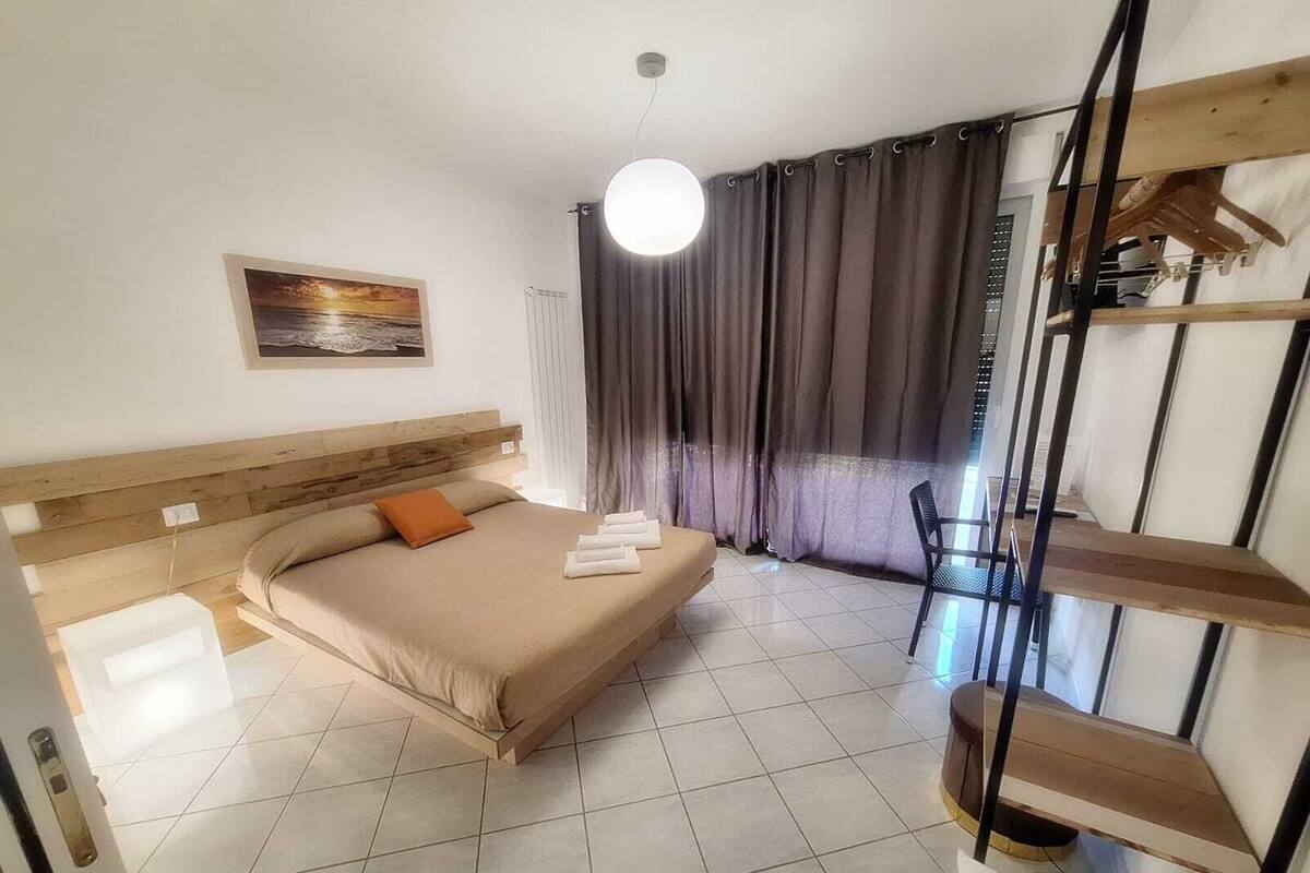 Ein Hotelzimmer mit Bett, einem Bild an der Wand, einem Fenster mit Vorhängen, einem Sessel und einem Regal.