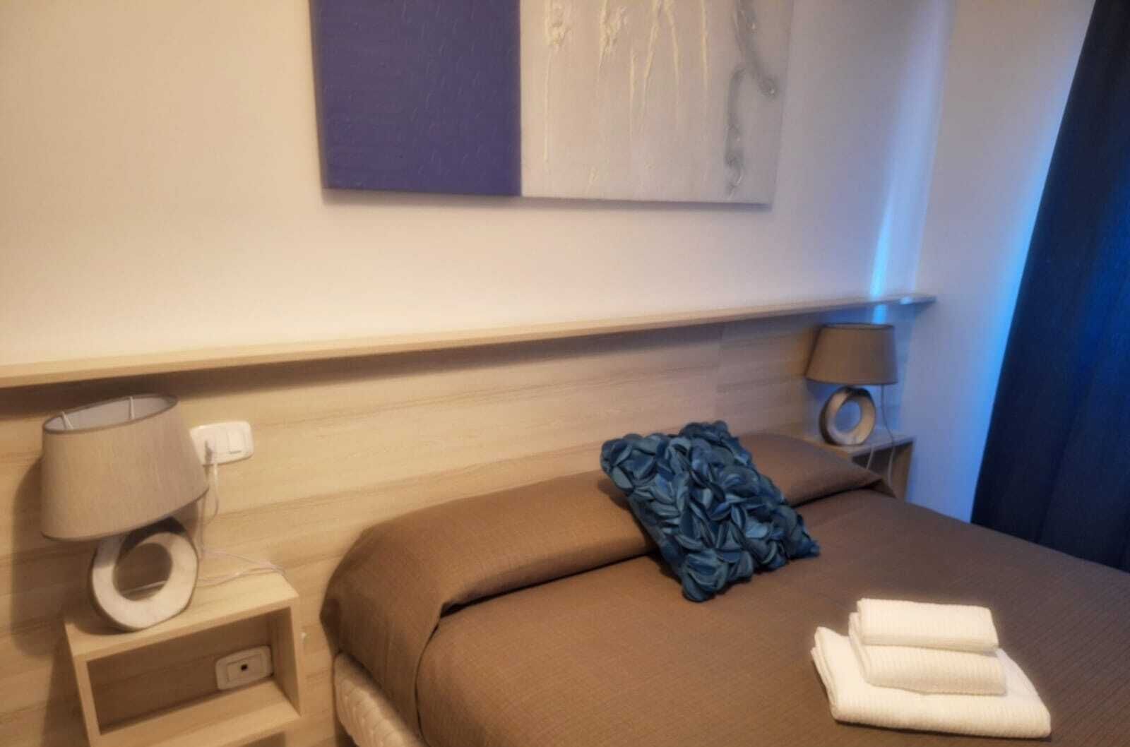 Una habitación de hotel con una cama, una mesita de noche con lámpara, una obra de arte en la pared y una ventana con cortinas.
