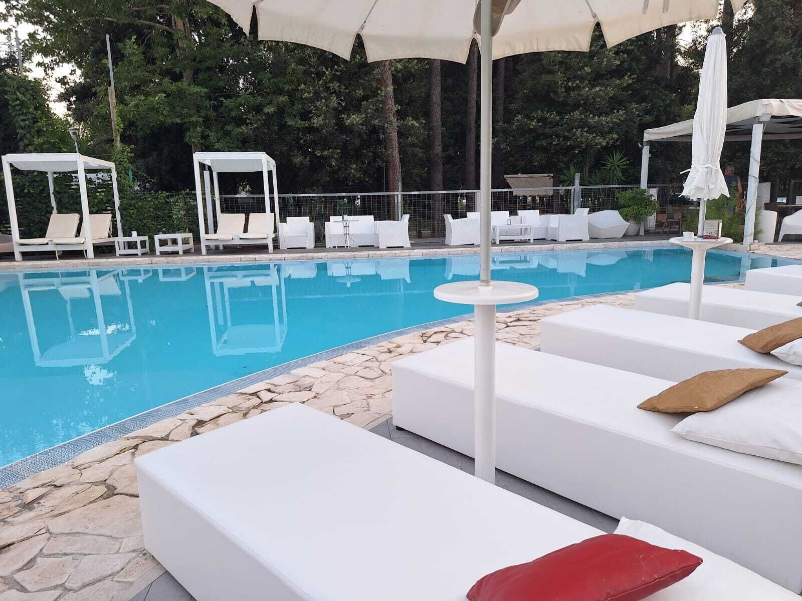 A poolside area with white lounge chairs and umbrellas, surrounded by greenery.