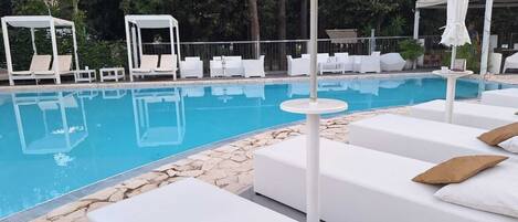 A poolside area with white lounge chairs and umbrellas, surrounded by greenery.