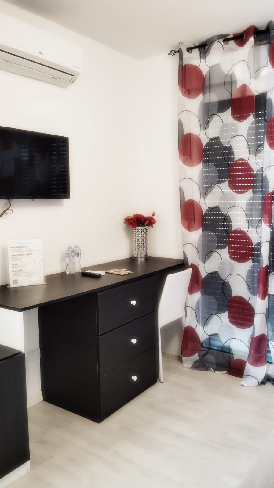 Un baño moderno con un tocador negro, un televisor montado, una cortina de ducha con estampado rojo y gris, y un florero con flores rojas en la encimera.