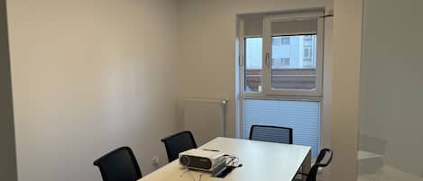 A conference room with a white table, four black chairs, a projector, and a window with blinds.