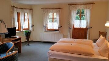 Ein Schlafzimmer mit einem Bett, einem Schreibtisch mit Computer, einem Sessel und zwei Fenstern mit Vorhängen.