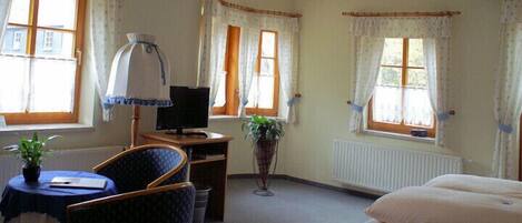 A room with a bed, a desk with a computer, a chair, a small table with a plant, and a radiator.