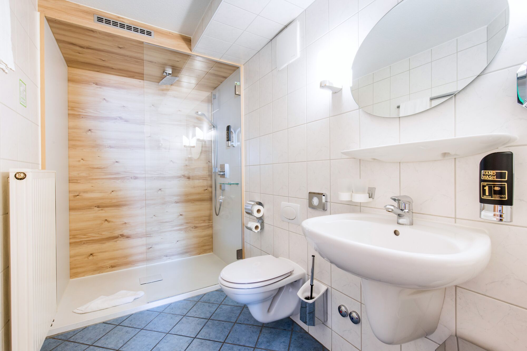 Un baño moderno con cabina de ducha de madera, inodoro blanco, lavamanos con dispensador de jabón y un espejo grande.