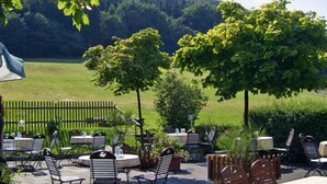 Een terras buiten met tafels en stoelen, omringd door groen en bomen.