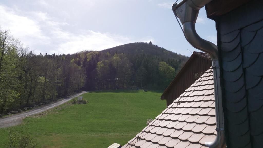 Vista de una casa con techo inclinado y chimenea, con vista a un campo verde y una colina boscosa.