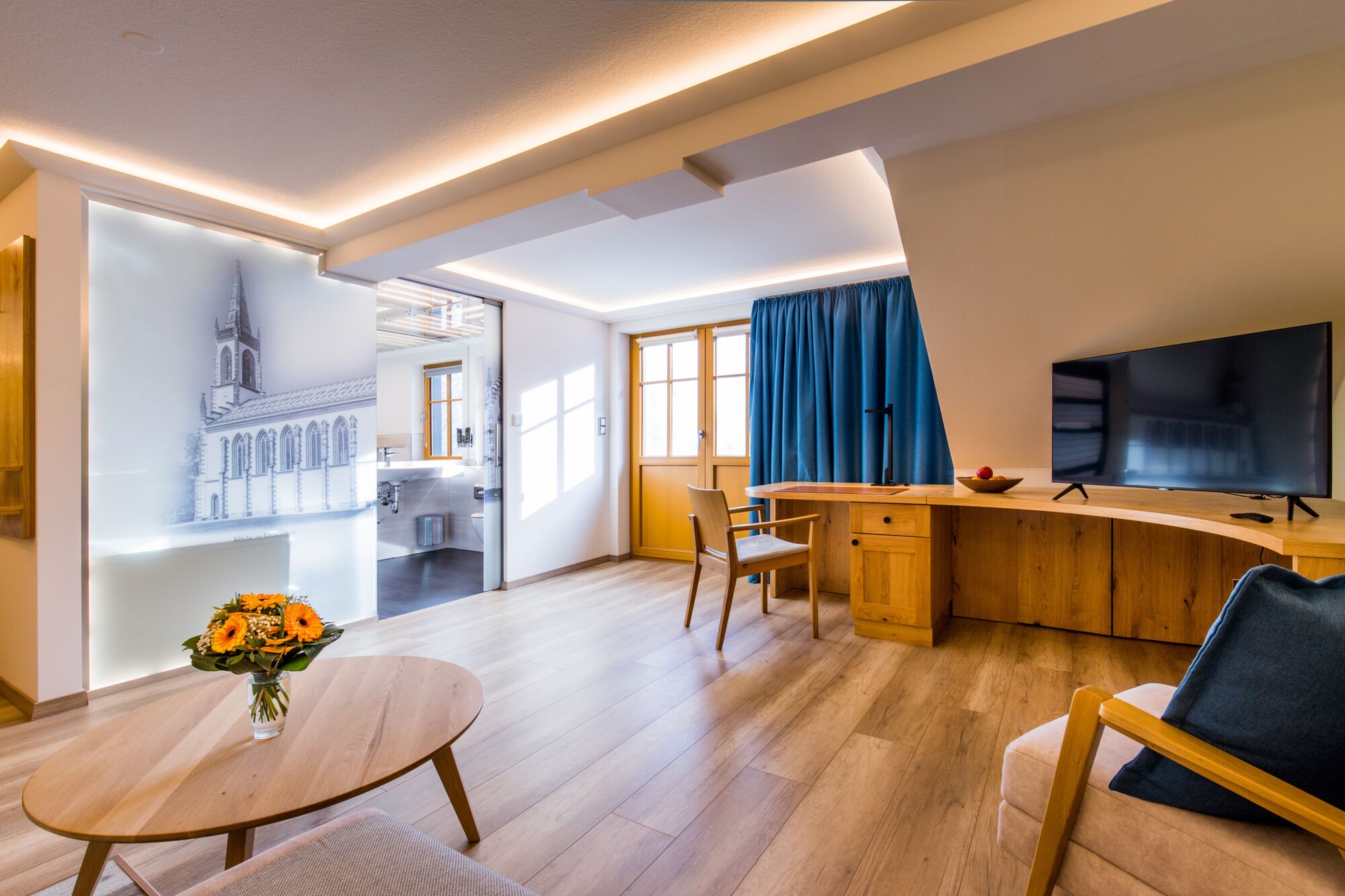 Een moderne woonkamer met houten vloer, een eettafel met stoelen, een televisie en een keuken die door een open deur zichtbaar is.