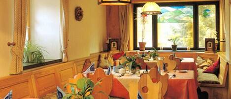 Comedor con mesas y sillas de madera, plantas en macetas y un gran ventanal con vistas a zonas verdes.
