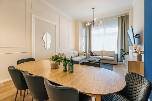 A modern living room with a round wooden dining table, dark upholstered chairs, a sofa, a coffee table, and a TV mounted on the wall.