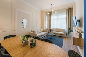 A modern living room with a round wooden table, sofas, a TV mounted on the wall, and a large window with curtains.