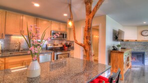 A modern kitchen with granite countertops, stainless steel appliances, and a wooden tree trunk centerpiece. - Killington Tree House: 2 Room Modern Condo 432 (Killington)