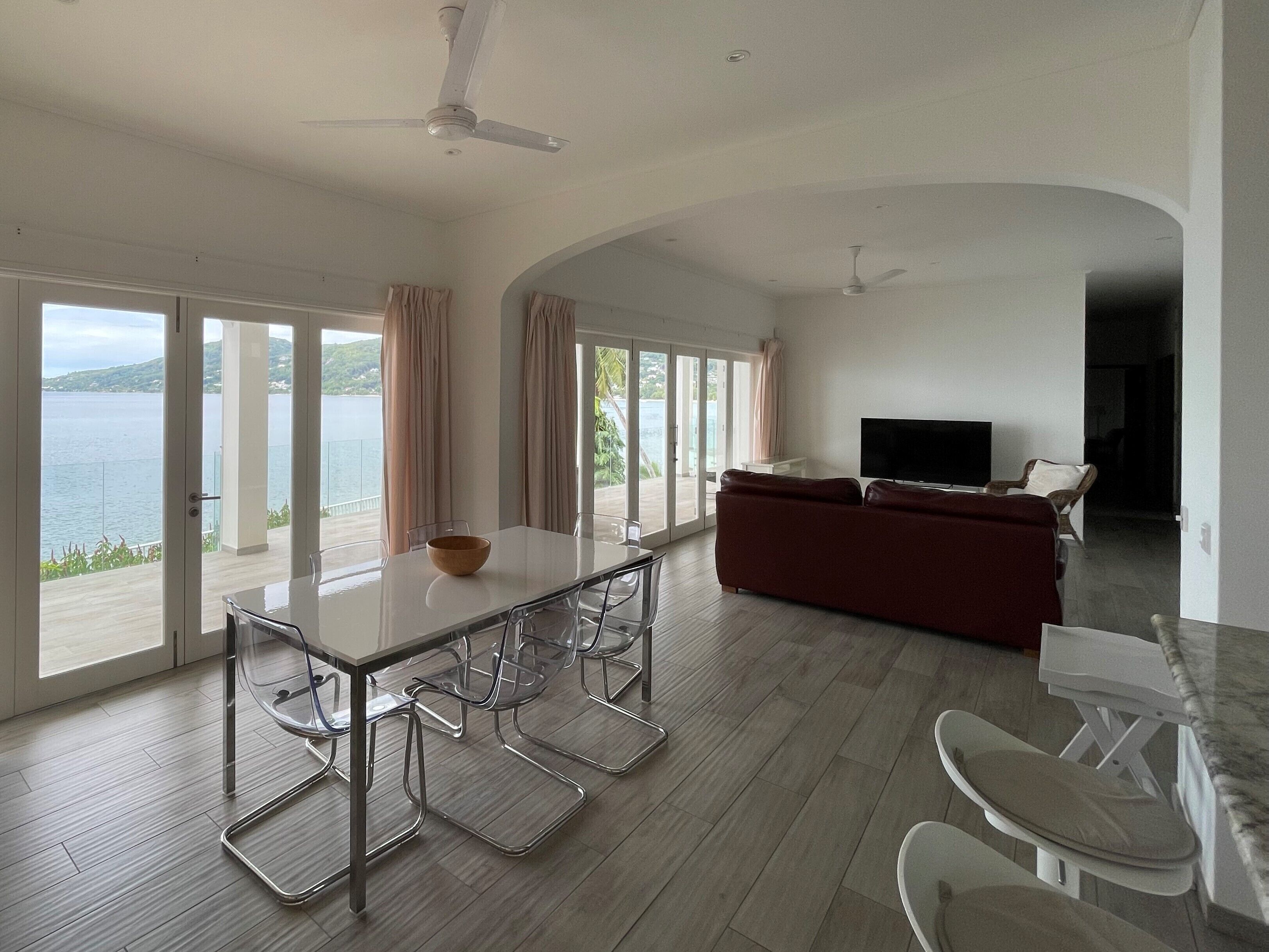 A spacious living area with a dining table, chairs, a sofa, and a TV. Large windows offer a view of the sea.