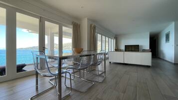 A modern living room with a glass dining table and chairs, a sofa, and a TV. Large windows offer a view of the sea.