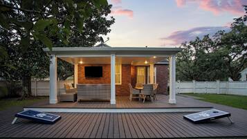 A covered patio with a brick wall, a flat-screen TV, wicker furniture, and two cornhole boards.