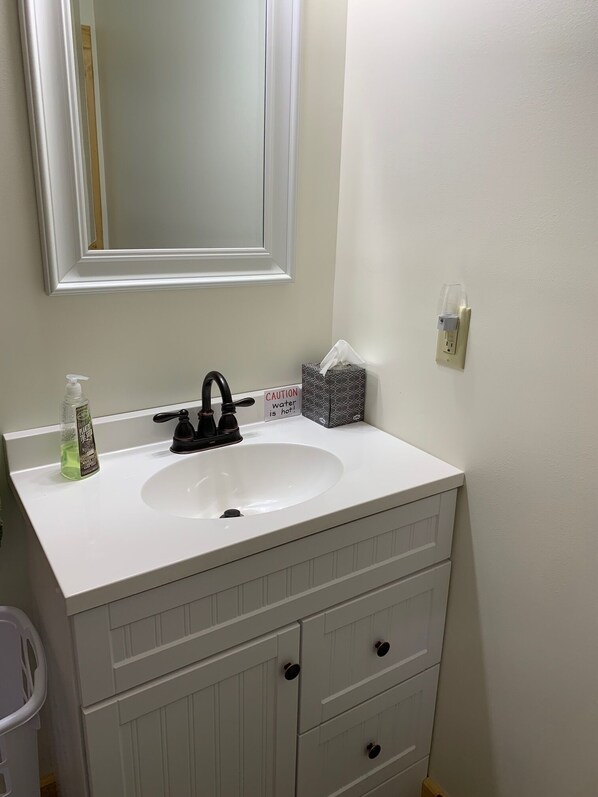 Une salle de bain avec un lavabo blanc, un robinet noir et un miroir au-dessus. On peut voir un panneau d’avertissement sur le mur, une boîte de mouchoirs et un distributeur de savon sur le lavabo.