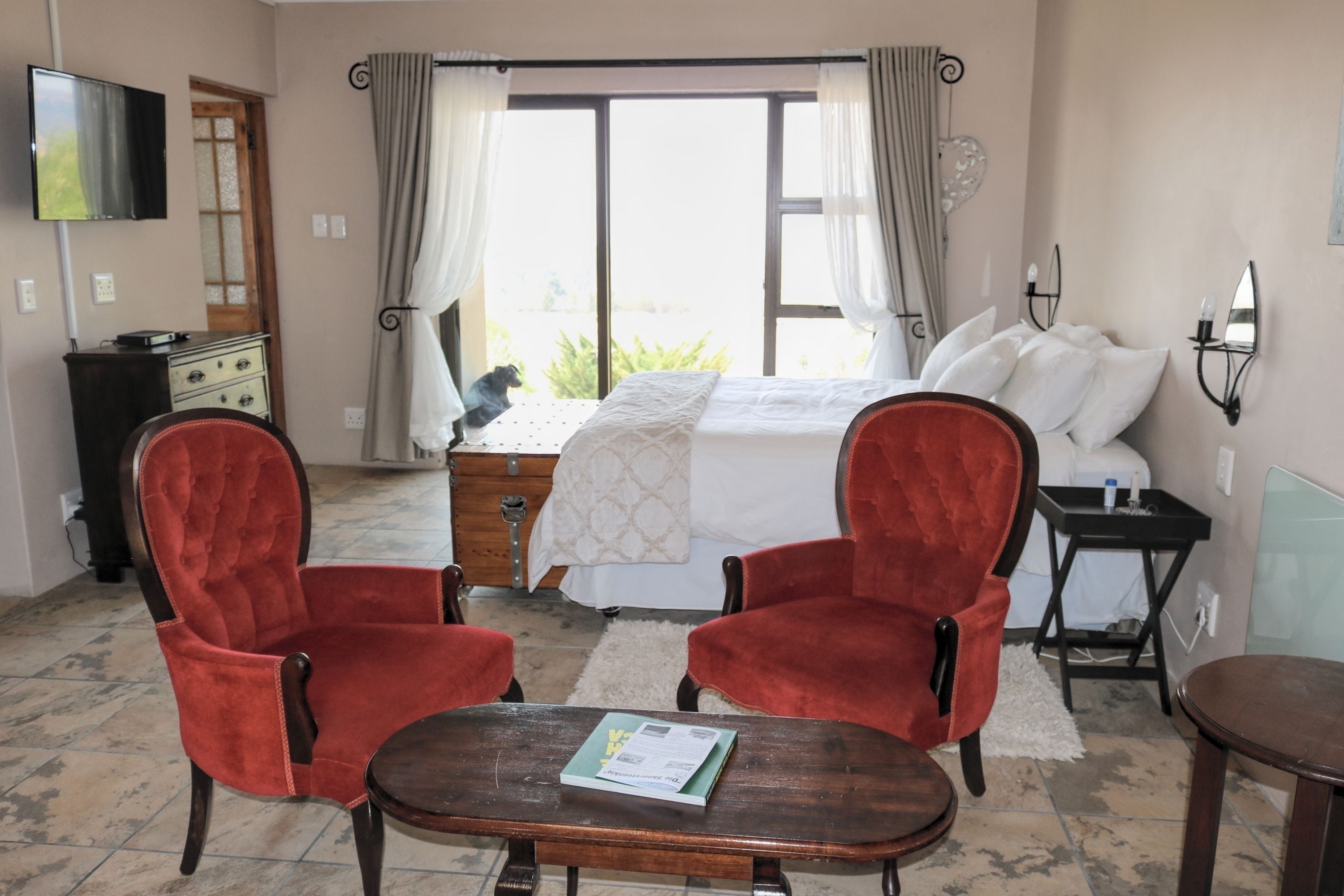 A hotel room with a bed, two red armchairs, a wooden table, a TV, and a window with curtains.