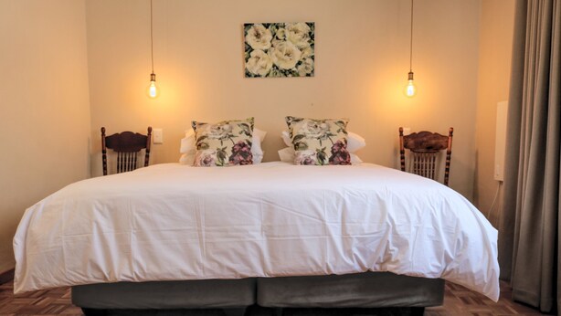 A large bed with a white duvet, two floral pillows, and a headboard. There are two wall-mounted light fixtures and a painting above the bed. - The Fat Mulberry Guesthouse (Fouriesburg)