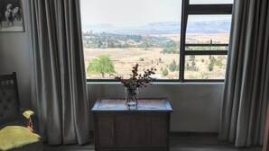 Una habitación con vistas a un paisaje rural a través de una ventana, una mesa pequeña con un florero de flores secas y una silla con cojín.