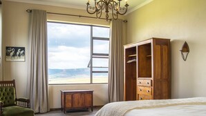 A bedroom with a large bed, a wooden wardrobe, a green armchair, a small table, and a view of the beach through a window. - The Fat Mulberry Guesthouse (Fouriesburg)