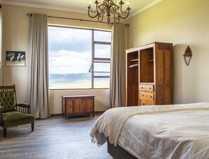 A bedroom with a large bed, a wooden wardrobe, a green armchair, a small table, and a view of the beach through a window. - The Fat Mulberry Guesthouse (Fouriesburg)