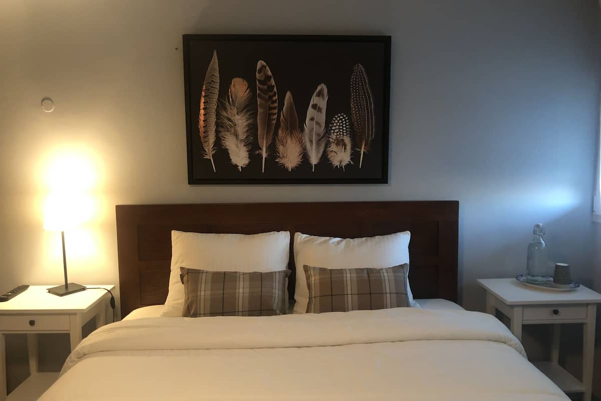 Een keurig opgemaakt bed met een hoofdeinde, twee nachtkastjes met lampen, en een lijst met een afbeelding van veerveren aan de muur.